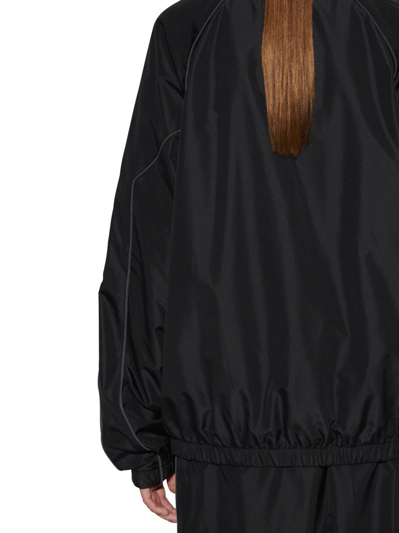 Person mit langen, glatten braunen Haaren, die eine schwarze, leichte Jacke und passende schwarze Hose trägt, von hinten gezeigt.