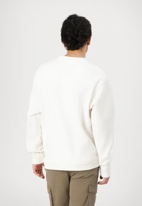 Hvit bomullssweatshirt med en myk tekstur, rund hals og ribbestrikkede mansjetter. Iført olivengrønne cargo-bukser, sett bakfra.