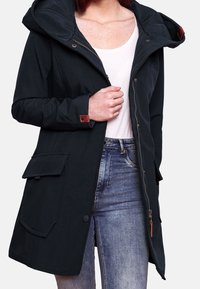 Svart lättviktig jacka med huva, dragkedja framtill, två stora fickor med lappar och bruna läderdetaljer på ärmarna. Slät yta.