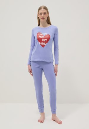 Vrouw staat op blote voeten en draagt een lichtpaarse top met lange mouwen en een broek, de top heeft een rood hart met Franse tekst "L'amour rend tout possible."