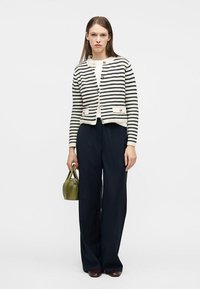 Γυναίκα με μαύρη και λευκή ριγωτή ζακέτα με κουμπιά, μαύρο παντελόνι με φαρδιά πόδια, κρατώντας μικρή πράσινη τσάντα, στέκεται μπροστά σε λευκό φόντο.