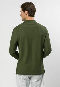 Olivovo zelená polokošeľa s dlhým rukávom, zaobleným golierom a rovnou spodnou časťou. Hladká textúra s bočnými rozparkami. Nosená s svetlými džínami.