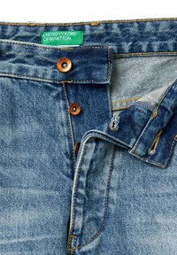 Jeans de mezclilla azul con el cierre parcialmente abrochado y una etiqueta verde que dice "United Colors of Benetton" en la cinturilla.