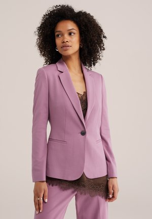Vrouw met krullend haar die een lavendelkleurige op maat gemaakte blazer en broek draagt, gelaagd over een bruin hemdje met kant.