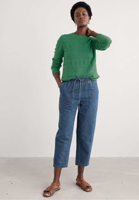 Zaļš adīts džemperis ar strukturētu rakstu, kombinēts ar vaļīgiem ziliem džinsa biksēm ar elastīgu jostasvietu un divām priekšējām kabatām. Brūnas čības.