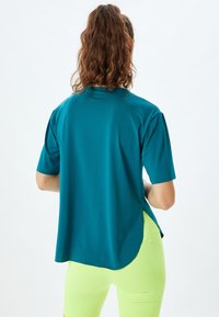 Teal kortærmet top med en afslappet pasform og sideslits, der har en glat tekstur. Pair med lysegule shorts.