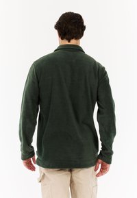 Muškarac u tamnozelenoj flis jakni i bež cargo hlačama, stoji okrenut leđima prema kameri na bijeloj pozadini.