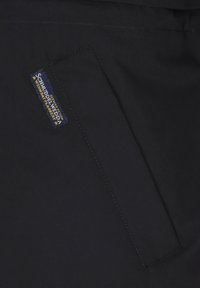 Sort stof med en rektangulær lomme, der har en label med teksten "Schmuddelwedda" i gul og blå syning. Glat tekstur.