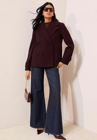 Donna che indossa un cappotto doppiopetto di colore prugna scuro, jeans a gamba larga scuri, tacchi a punta, occhiali da sole e porta una piccola borsa abbinata.