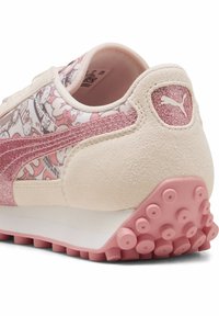 Le sneakers PUMA presentano una tomaia in suede rosa chiaro con dettagli in glitter e un motivo a cartoni animati, una suola in gomma con borchie rosa in rilievo e una finitura liscia.