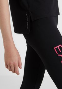 Schwarzes Baumwoll-T-Shirt mit einem leicht verlängerten Saum und seitlichem Schlitz. Schwarze Leggings mit großen rosa Text auf dem Oberschenkel.