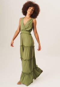 Olive green maxi dress with a V-neckline, smocked waist, tiered design, and ruffled accents. Made from lightweight fabric, suitable for warm weather.