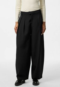 Czarne spodnie o szerokich nogawkach z plisami, wysokim stanem i gładką fakturą materiału. Połączone z błyszczącymi czarnymi butami.