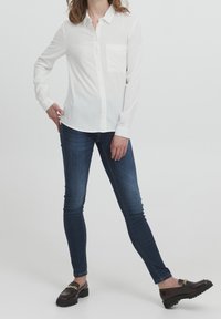 Chemise blanche à manches longues avec poche poitrine, associée à un jean slim bleu foncé et des chaussures sombres ornées d'une chaîne dorée.