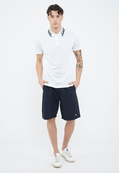 Young man standing with hands in pockets wearing white polo shirt, navy shorts, white sneakers, and visible tattoos on right arm.