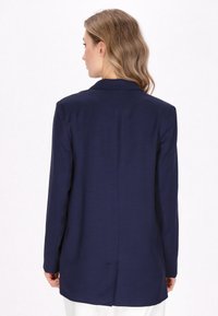 Navy modrý blazer s hladkým povrchem, dlouhými rukávy a klasickým límcem. Obsahuje rozparek na zadní straně a otevřený přední design.