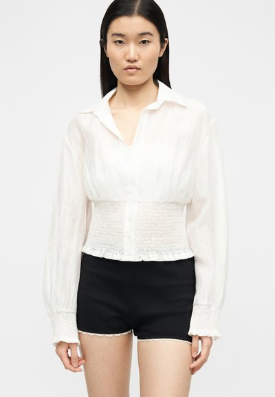 Femme portant un chemisier blanc à manches longues avec taille et poignets smockés, associé à un short taille haute noir avec bordure en dentelle.