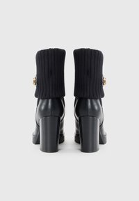 Black high-heeled boots with ribbed black knit fold-over cuff, smooth leather upper, chunky heel, and gold accent detail on the side.