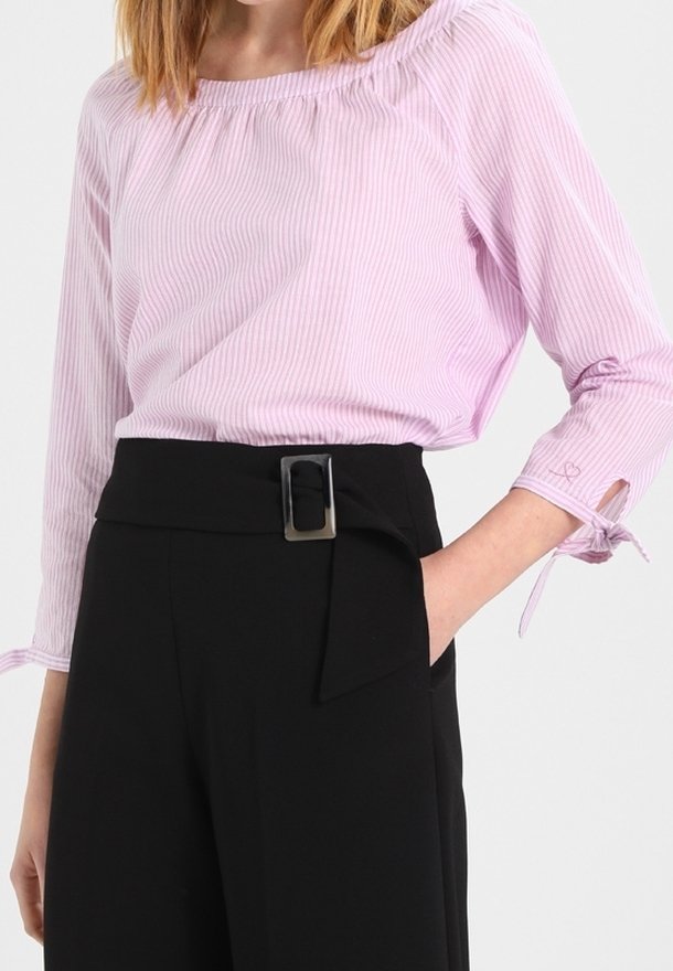 Femme portant un chemisier à manches longues rose clair avec des rayures discrètes, rentré dans un pantalon noir taille haute avec une large ceinture et une boucle rectangulaire.