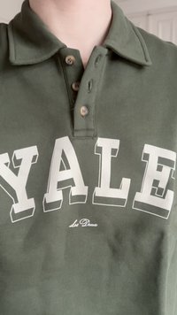 Asmuo vilkintis alyvuogių žalios spalvos marškinėlius su apykakle, rudais mygtukais ir dideliu baltu užrašu "YALE" ant krūtinės.