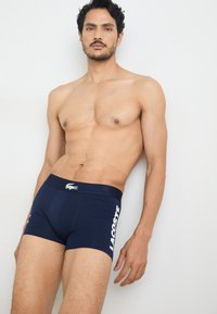 Navyblauwe katoenen boxershorts met een strakke pasvorm, voorzien van een wit "LACOSTE" logo aan de zijkant en een klein krokodillenembleem op de tailleband.