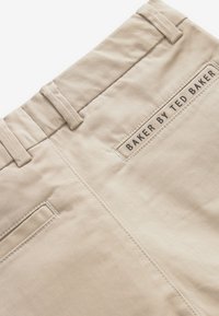 Pantalon beige avec passants de ceinture et poches arrière cousues, avec une étiquette "Baker by Ted Baker" au-dessus de la poche droite.