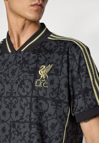 Fekete mintás futballmez arany Liverpool FC logóval, sárga részletekkel a V-nyakú galléron és az ujjakon.