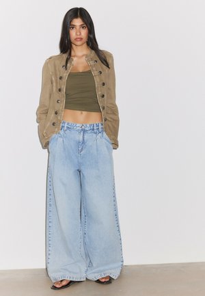 Kvinde med langt mørkt hår iført olivengrøn crop top, tanfarvet knappet jakke, vide lyseblå jeans og sorte sandaler, står mod en ensfarvet væg.