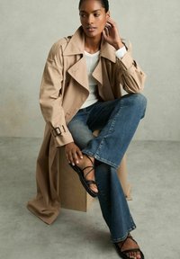 Beige trench coat dengan desain klasik, dipakai di atas atasan lengan panjang putih dan celana jeans flare biru, dipadukan dengan sandal bertali hitam.