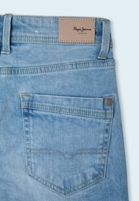 Svetle modré džíny z denimu s koženou nášivkou, detaily šití na zadních kapsách a viditelným švem podél švů.