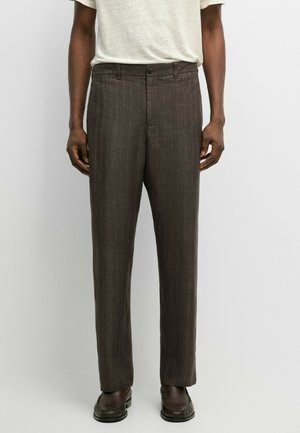 Uomo che indossa pantaloni a righe marrone scuro con chiusura a bottone e cerniera, abbinati a scarpe marrone scuro e camicia chiara.