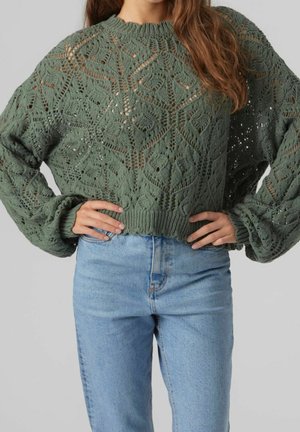 Femme portant un pull vert tricoté avec des motifs de feuilles et un jean taille haute bleu clair, les mains posées sur les hanches.