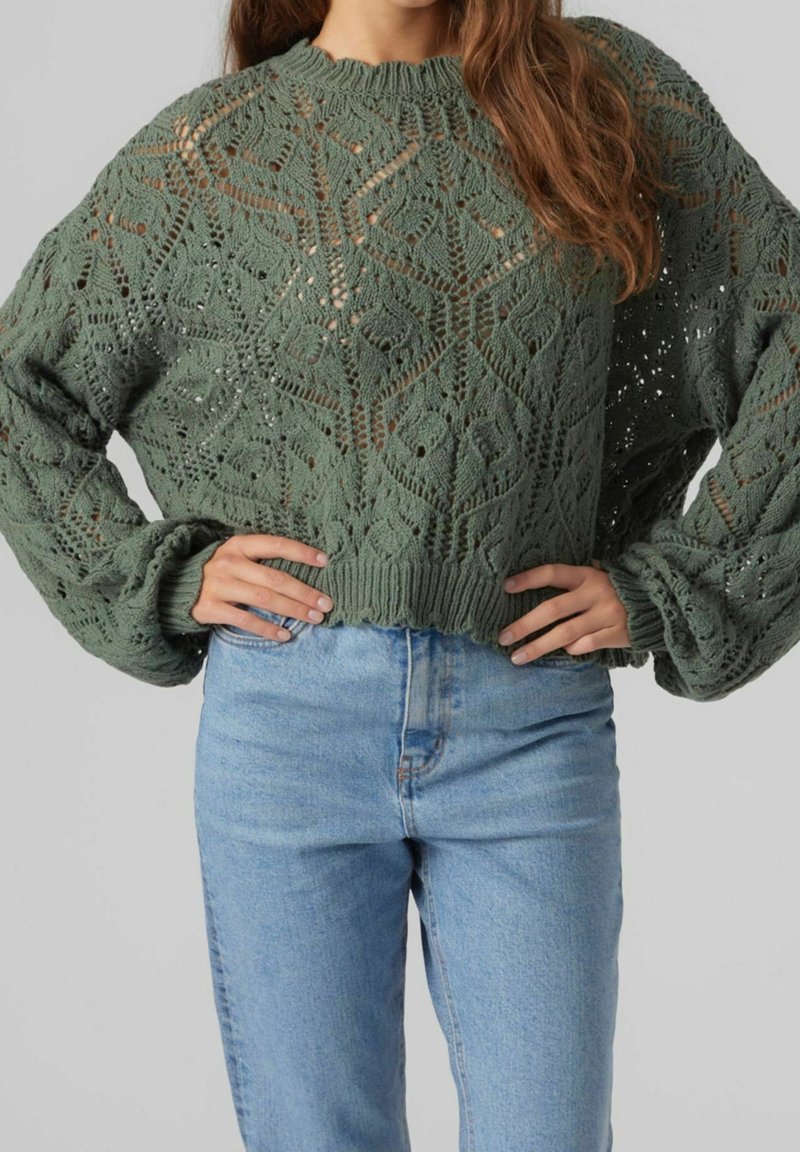 Femme portant un pull vert tricoté avec des motifs de feuilles et un jean taille haute bleu clair, les mains posées sur les hanches.