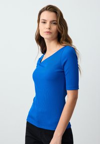 Top blu ribbed con un collo asimmetrico unico e maniche corte. Il tessuto appare liscio con un design aderente.
