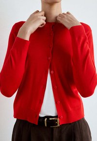 Personne boutonnant un cardigan rouge vif sur une chemise blanche, portant un pantalon foncé avec une ceinture noire à boucle dorée.