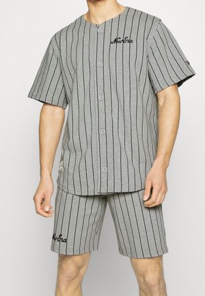 Mann trägt graues Hemd mit Knöpfen und Shorts mit schwarzen vertikalen Streifen und "New Era" Logo auf Brust und Oberschenkel.
