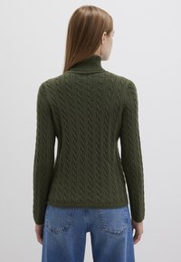 Dunkelgrüner, kabelgestrickter Pullover mit hohem Kragen und langen Ärmeln, ausgestattet mit einem gerippten Saum und einem markanten Texturmuster, kombiniert mit blauen Jeans.