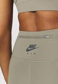 Sportovní oblečení Nike s hladkým olivově zeleným materiálem. Obsahuje viditelné logo značky a malou kapsu na zip na pase.