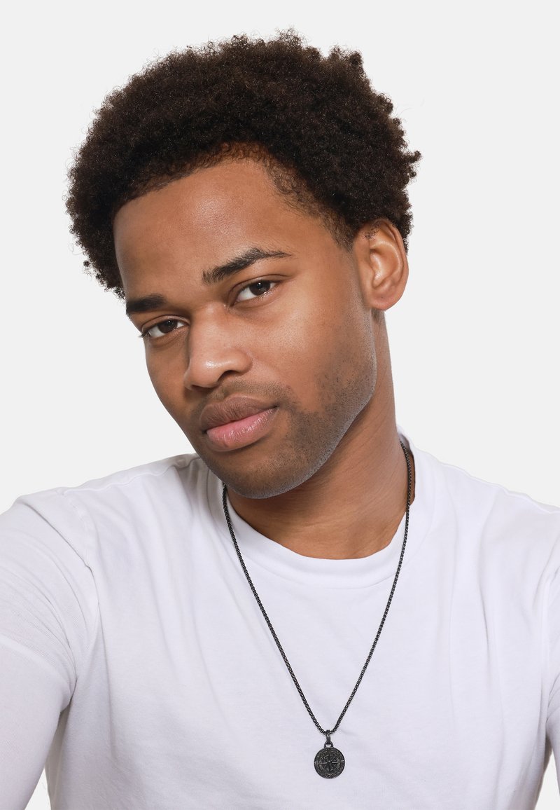 Zwarte hangerketting op een metalen ketting gedragen over een wit T-shirt. Onderwerp heeft kort, krullend haar en een neutrale uitdrukking.