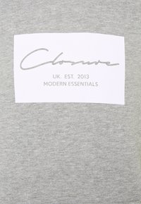 Tissu gris avec une étiquette rectangulaire blanche indiquant « Closure » en cursive, et en dessous « UK. EST. 2013 MODERN ESSENTIALS » en majuscules.