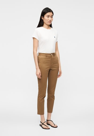 Valge T-särk väikese sinise logoga, pruunid kitsad püksid ja mustad madalad sandaalid. Sile kangas, kitsa lõikega püksid ja üles keeratud varrukaääred.