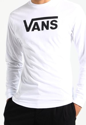 Biała koszula z długim rękawem z czarnym logo "VANS" na piersi, noszona przez osobę z rękami w kieszeniach czarnych spodni.