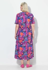 Maxikleid in leuchtendem Lila mit floralen und blattartigen Mustern in Pink, Orange und Türkis. Kurze Ärmel und tailliert geschnitten. Dazu pinke Schuhe kombiniert.