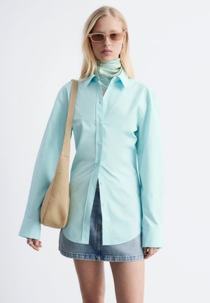 Blondynka w jasnoniebieskiej koszuli zapinanej na guziki, dżinsowej mini spódniczce, wzorzystej apaszce na szyję, brązowych okularach przeciwsłonecznych i z beżową torebką na ramieniu.