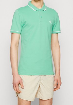 Buste d'homme portant un polo à manches courtes vert menthe avec un liseré blanc et un short beige sur un fond uni.
