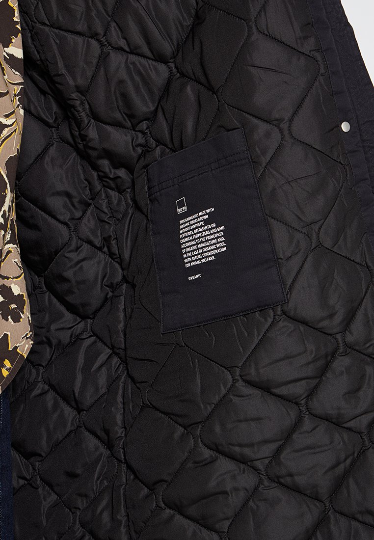 Gevoerde zwarte jas met een patch pocket waarin zorginstructies zijn opgenomen. De stof heeft een gestructureerd oppervlak en een gladde afwerking.