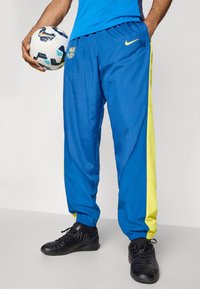 Niebieskie sportowe spodnie z żółtymi bocznymi panelami, elastycznym pasem i logo Nike. Gracz trzyma wzorzystą piłkę nożną w jednej ręce.