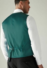 Green satin vest with a smooth texture, adjustable back strap, worn over a white long-sleeve shirt, viewed from the rear.
