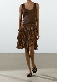 Haut marron sans manches associé à une jupe marron à volants avec motif, la superposition crée une apparence fluide, complétée par des tongs noires.