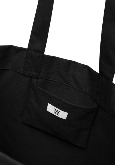 Schwarze Canvas-Umhängetasche mit zwei stabilen Griffen, ausgestattet mit einer kleinen Außentasche und einem weißen Etikett mit schwarzer Schrift. Einfaches, funktionales Design.